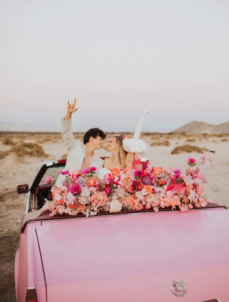 In questa foto due sposi si guardano e sorridono tenendo le braccia alzata mentre sono seduti a bordo di un'auto decappottabile di colore rosa e decorata con fiori di colore rosa e fucsia