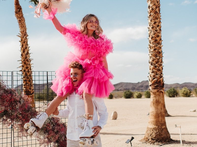 In questa foto due sposi di un matrimonio stile Barbie. La sposa sulle spalle dello sposo indossa un abito di tulle di colore fucsia e un paio di pattini a rotelle