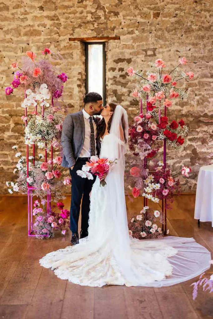 In questa foto due sposi si baciano davanti ad un backdrop floreale con fiori di colore rosa, fucsia, bianco per un matrimonio stile Barbie