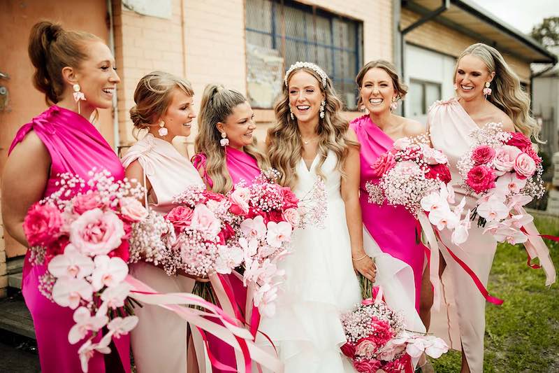 In questa foto una sposa sorridente è circondata dalle sue damigelle che indossano abiti monospalla di colore rosa e fucsia e tengono tra le mani bouquet abbinati