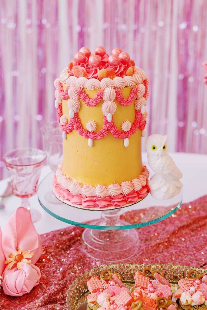 In questa foto una Lambeth Cake di colore giallo con riccioli di crema di colore rosa e fucsia e ciliegine decorate con glitter di colore rosa