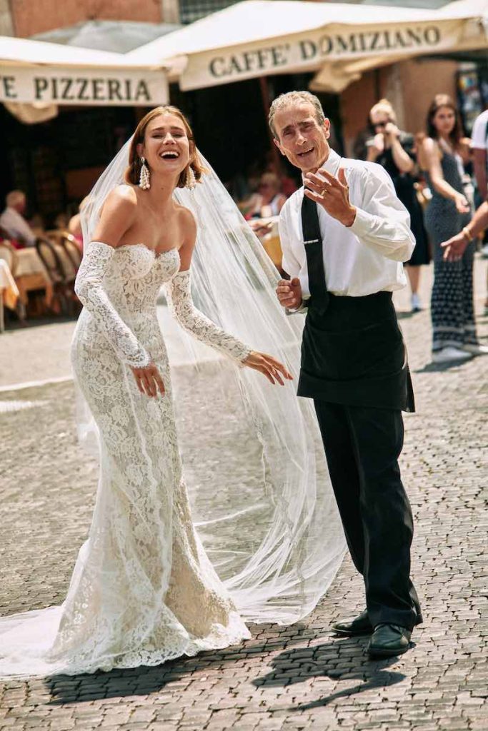 In questa foto una modella posa per le strade di Roma con un passante indossando un abito a sirena di Atelier Emé interamente in pizzo con velo lungo abbinato, scollo a cuore e maniche a guanto staccabili