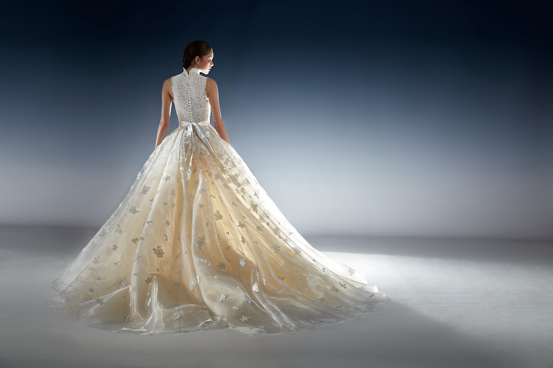 In questa foto una modella posa di spalle indossando un abito da sposa modello principessa della collezione esclusiva di Peter Langner con corpetto in pizzo floreale chiuso da un fiocco in raso e una gonna ampia colore avorio con fiori 3D all over