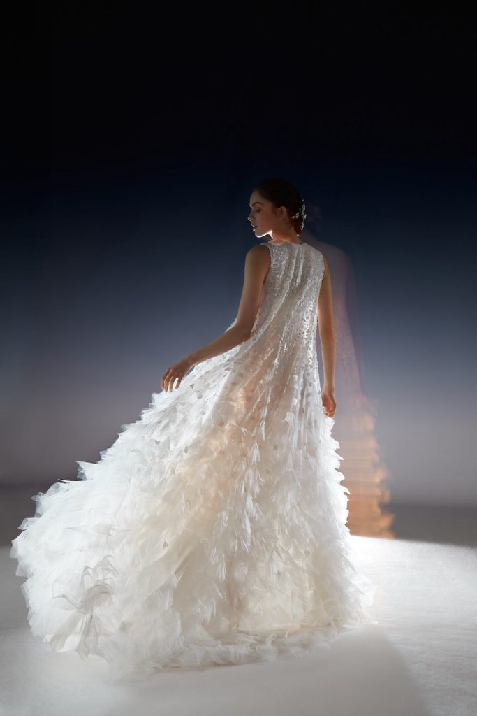 In questa foto una modella posa di spalle un abito da sposa della collezione White Gold 2024 di Peter Langner con petali 3D e ricami floreali sul corpetto