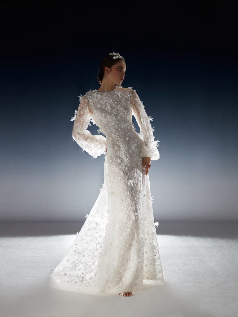 In questa foto una modella posa  indossando un abito da sposa della collezione White Gold 2024 di Peter Langner con maniche lunghe a sbuffo e petali e piume 3D