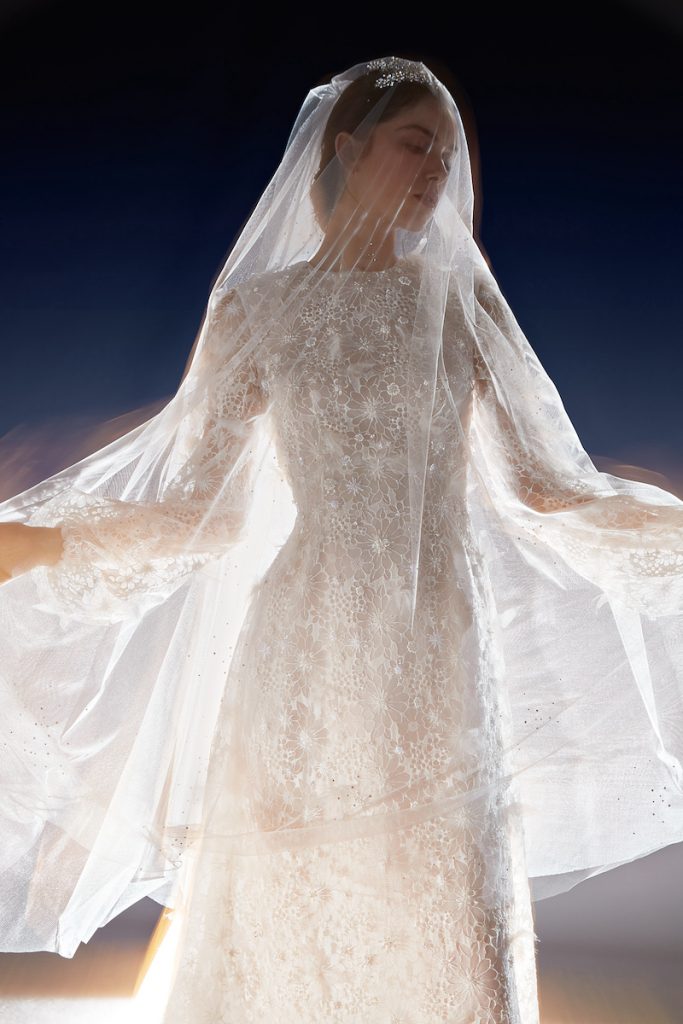 In questa foto una modella posa a mezzo busto indossando un abito da sposa ad A della collezione White Gold 2024 di Peter Langner con maniche lunghe e interamente in pizzo floreale. La modella indossa anche un velo in tulle a taglio vivo