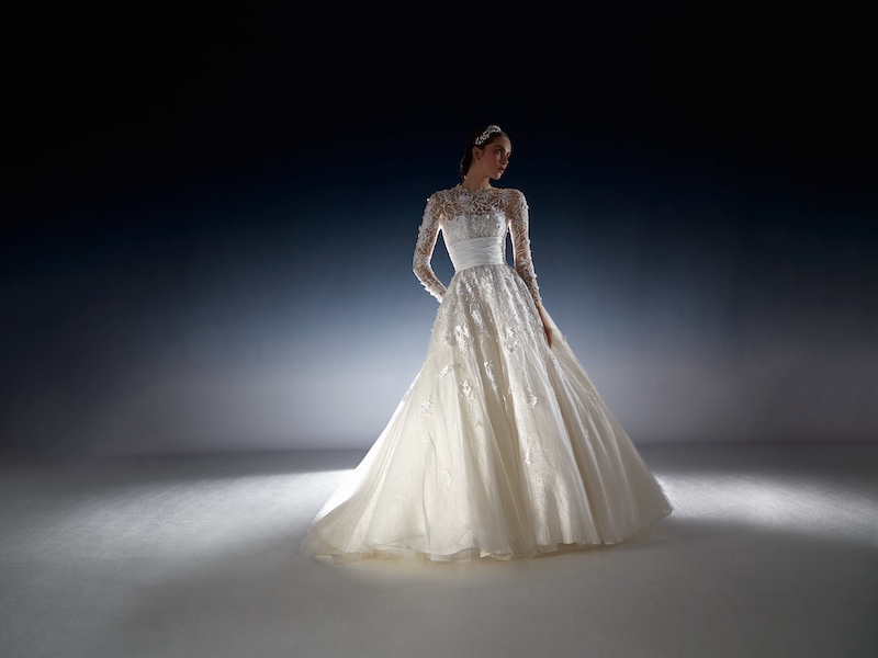 In questa foto una modella posa indossando un abito da sposa della collezione White Gold 2024 di Peter Langner in taffetà con corpetto a maniche lunghe ricamato con cintura in vita