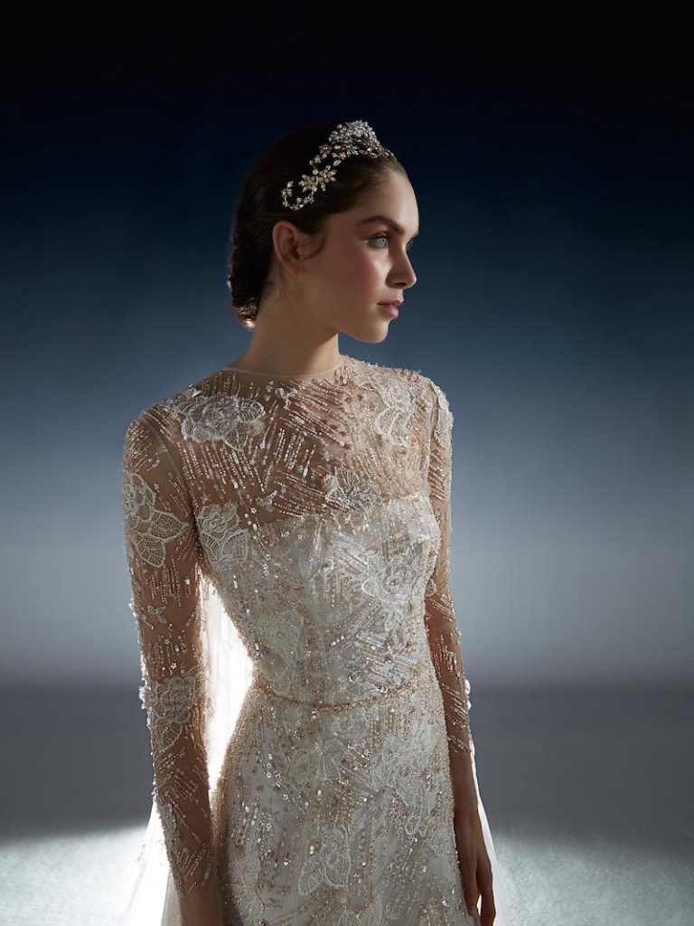 In questa foto una modella inquadrata a mezzo busto posa indossando un abito da sposa della collezione di lusso 2024 di Peter Langner con ricami di perline e pizzo colore bianco e oro 