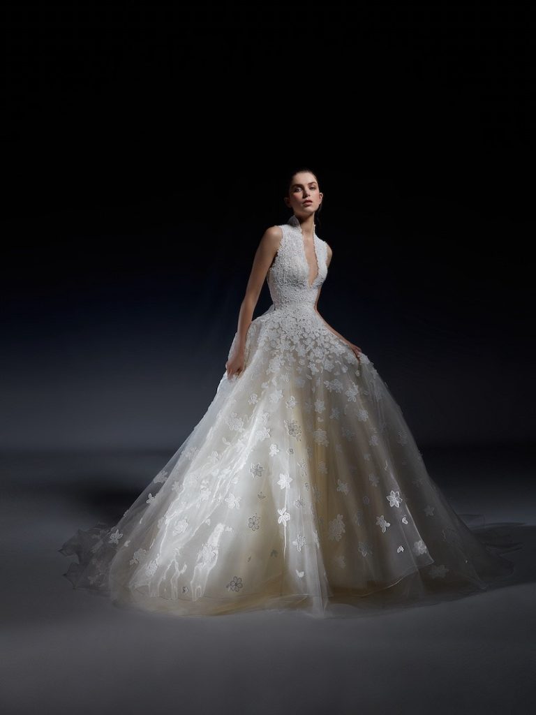 In questa foto una modella posa indossando un abito da sposa della collezione White Gold di Peter Langner modello a trapezio fiori 3D sul corpetto e sulla gonna e un profondo scollo a V