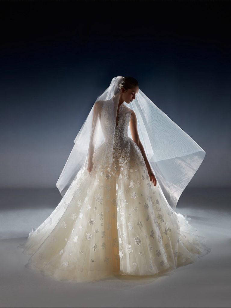 In questa foto una modella posa indossando un abito da sposa della collezione White Gold 2024 di Peter Langner modello ad A con ricami floreali all over e scollo Queen Anne. La modella indossa un velo a taglio vivo