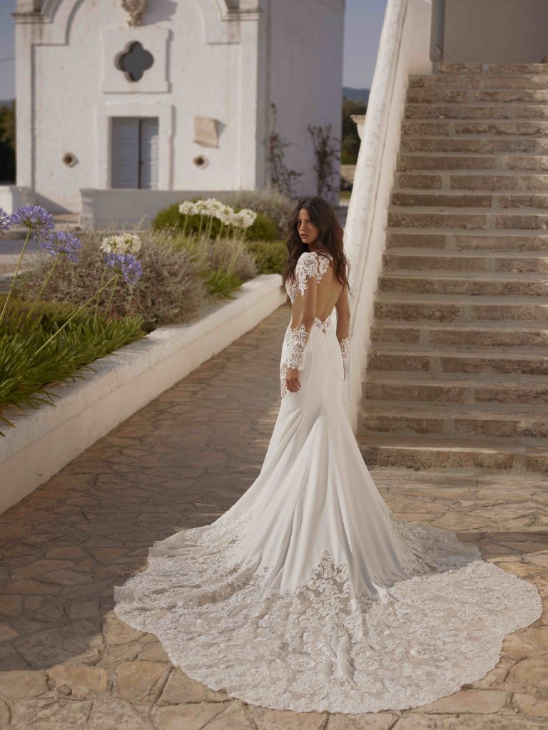In questa immagine un abito da sposa Capri 2024 con corpetto in pizzo e lunga coda ricamata.
