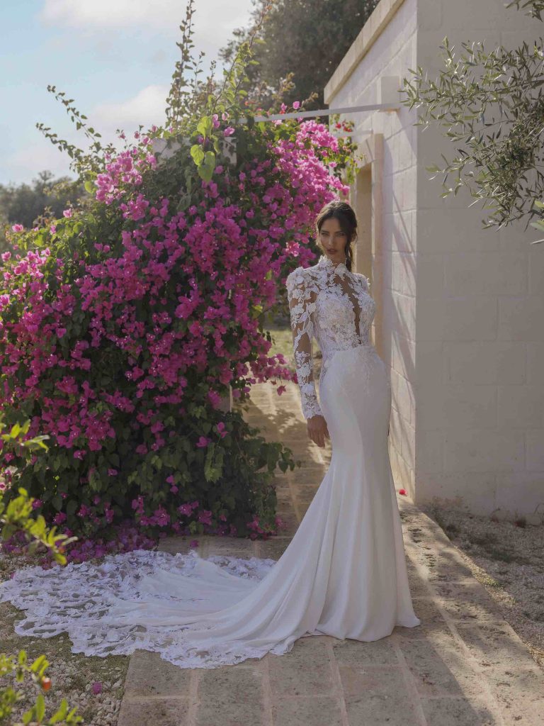 In questa immagine un abito da sposa Capri 2024 con corpetto in pizzo e lunga coda ricamata.