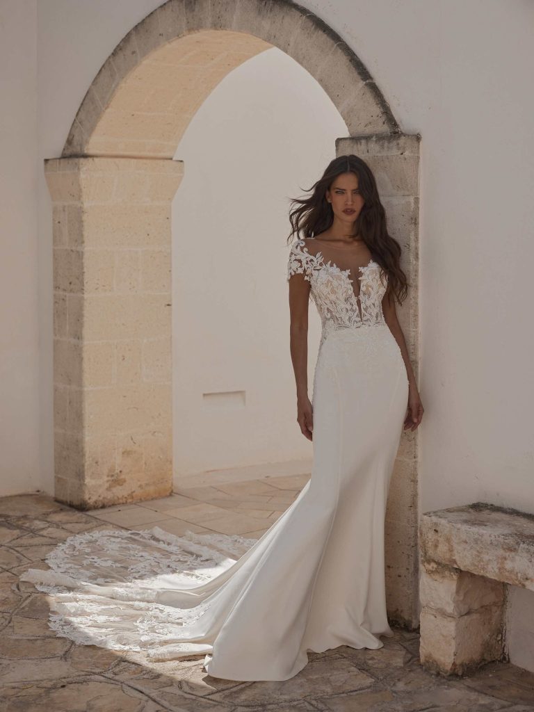 In questa immagine un abito da sposa Capri 2024 con corpetto in pizzo e lunga coda ricamata.
