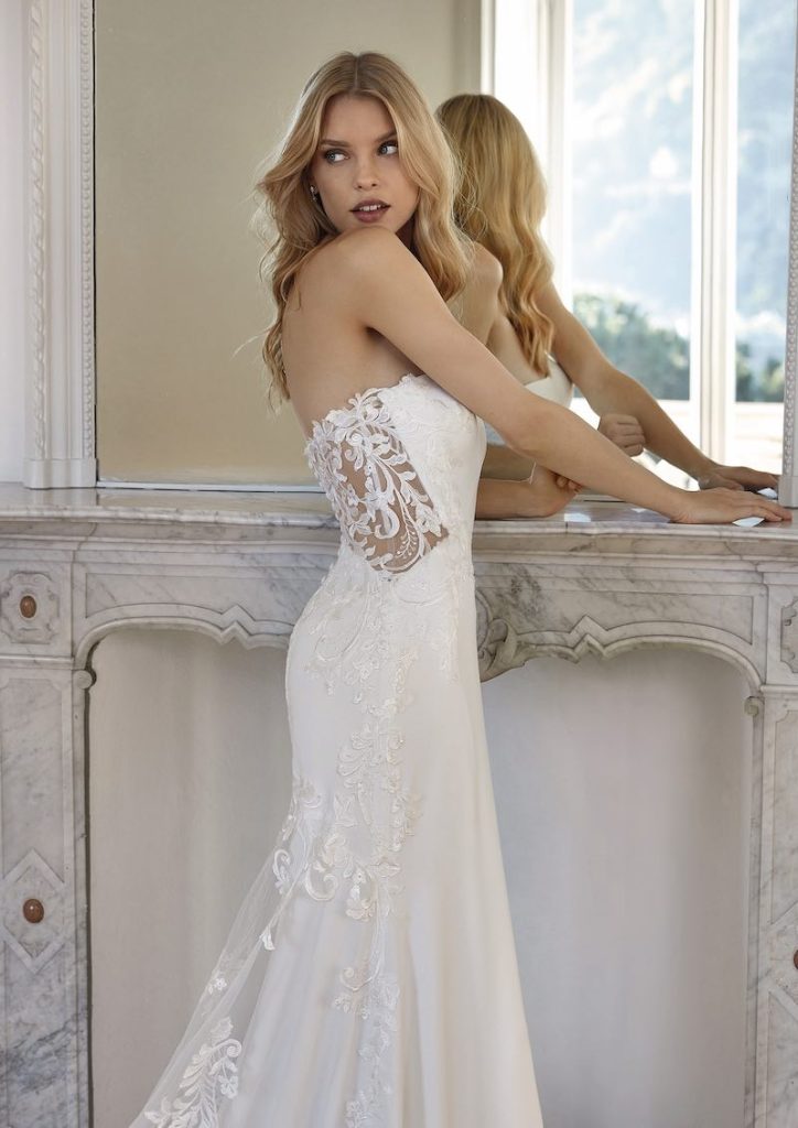 In questa foto una modella posa di tre quarti indossando un abito da sposa a sirena Nicole Milano 2024 in crêpe georgette con motivi jacquard di pizzo rebrodè sul retro