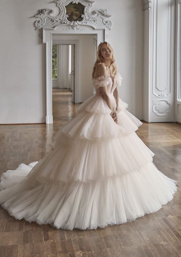 In questa foto una modella posa indossando uno degli abiti da sposa Nicole Milano 2024 modello principessa di colore rosa cipria con gonna di tulle a maxi balze
