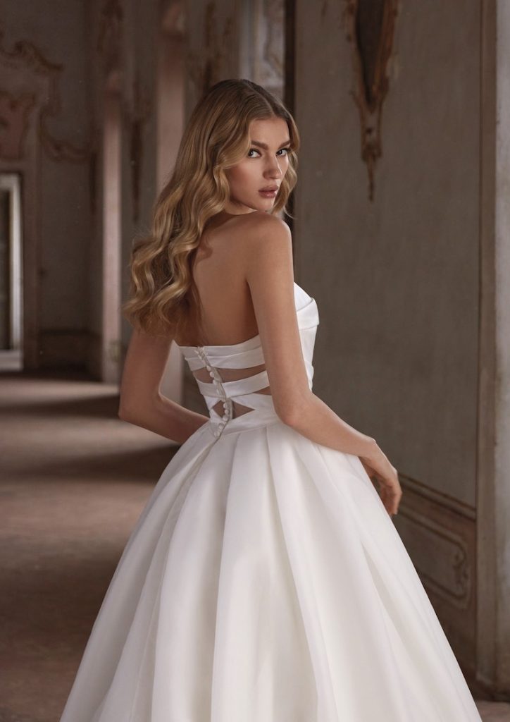 In questa foto una modella posa di spalle mostrando il dettaglio cut-out geometrico sulla schiena in un abito da sposa principessa Nicole Milano 2024
