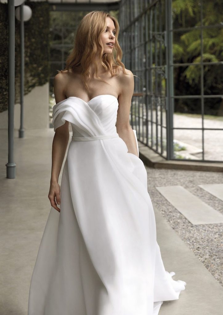 In questa foto una modella posa indossando un abito da sposa Nicole Milano 2024 modello ad A in chiffon con corpetto drappeggiato e scollatura a cuore
