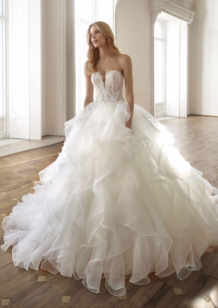 In questa foto una modella posa indossando un abito da sposa principessa Nicole Milano con corpetto senza spalline e scollo a cuore con motivo jacquard e un gonna con multi ruches in organza