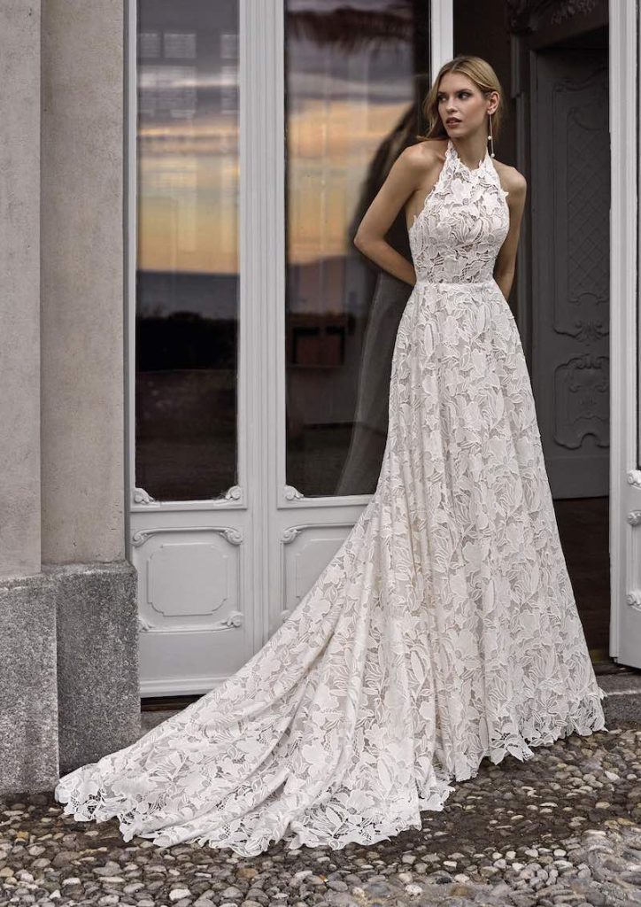 In questa foto una modella posa indossando un abito da sposa scivolato Nicole Milano 2024 in pizzo macramè con scollatura all'americana