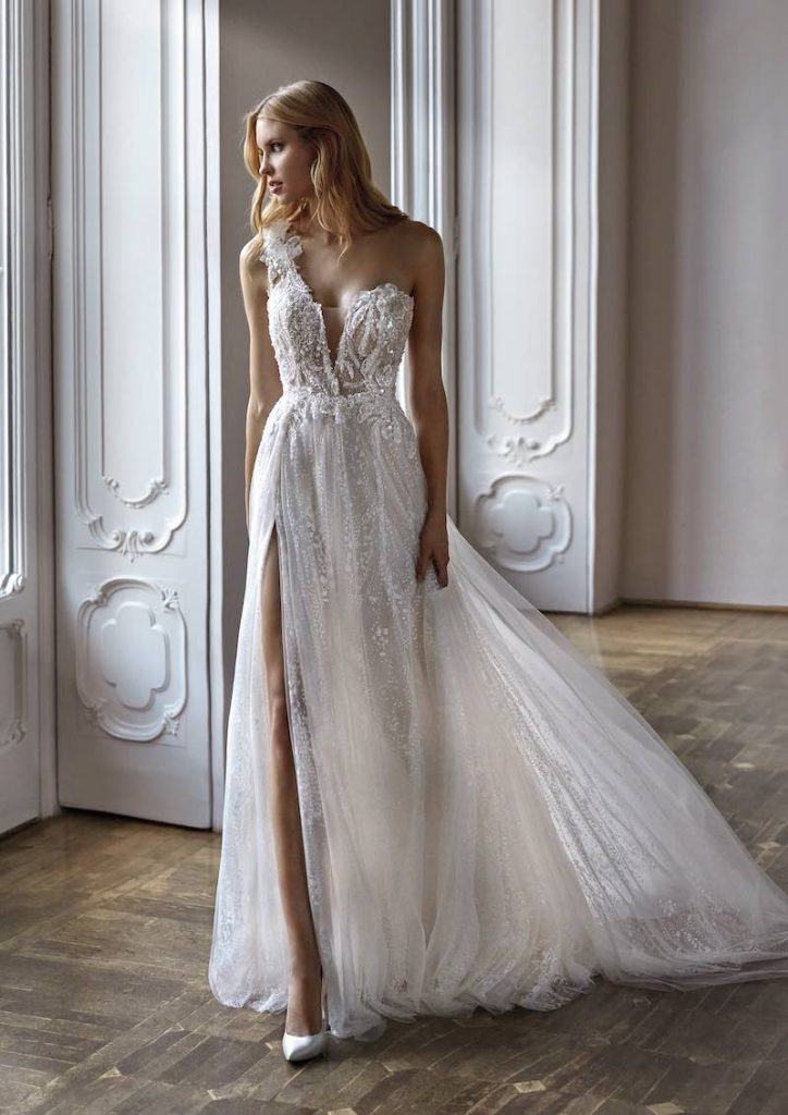 In questa foto una modella posa indossando un abito da sposa Nicole Milano 2024 modello scivolato in tulle sparkling con spacco laterale, scollatura monospalla e ricami di perline all over