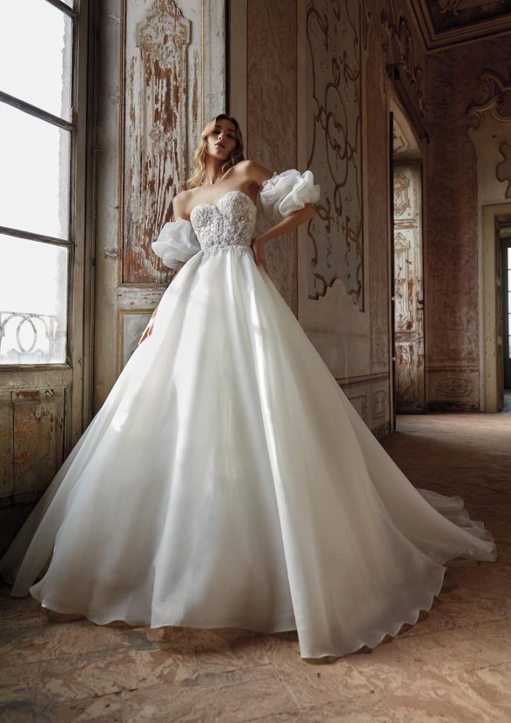 In questa foto una modella posa in un antico palazzo indossando uno degli abiti da sposa Nicole Milano 2024 modello ad A con gonna in organza, corpetto ricamato e maniche balloon staccabili