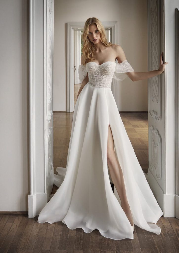 In questa foto una modella posa indossando uno degli abiti da sposa Nicole Milano 2024 modello scivolato con gonna in chiffon, spacco laterale e bustier con scollo a cuore ricamato con perline