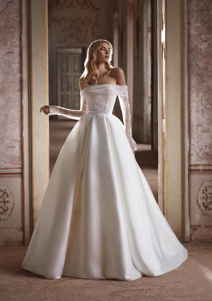 In questa foto una modella posa indossando un abito da sposa Redingote Nicole Milano con scollatura dritta, corpetto ricamato con cristalli, maniche lunghe a guanto 