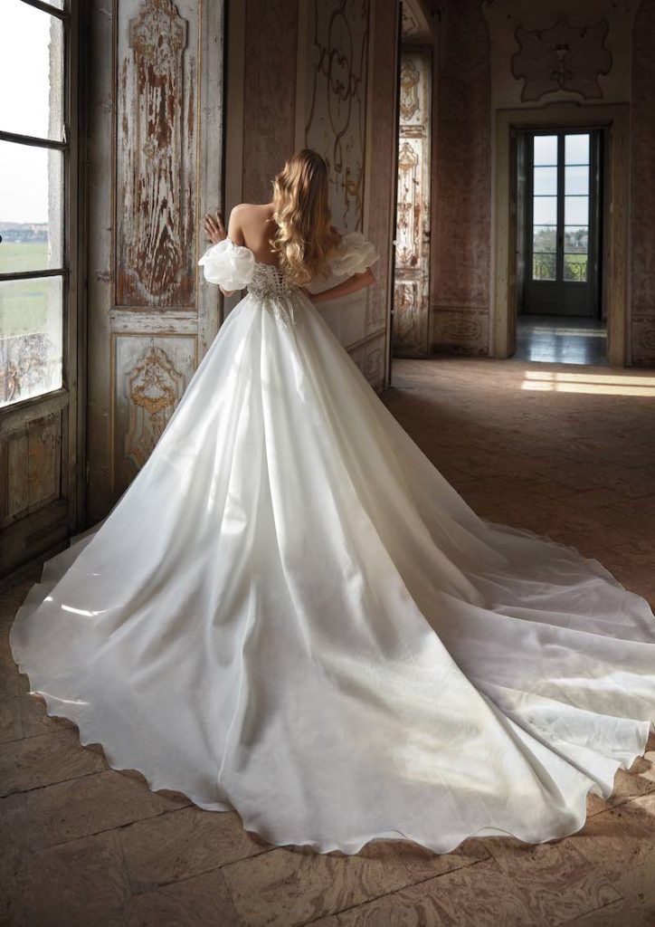 In questa foto una modella posa di spalle in un antico palazzo indossando un abito da sposa Nicole Milano 2024 con lunghissimo strascico in organza