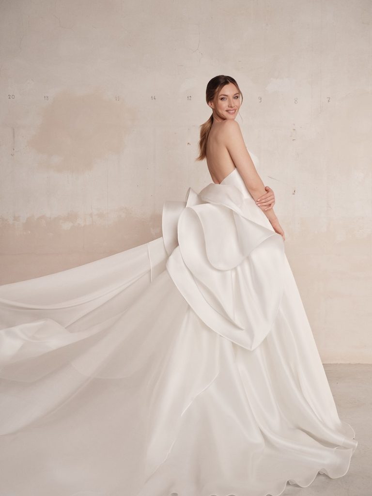 In questa foto una modella posa indossando una abito da sposa 2024 di Maria Pia Creazioni con gonna molto ampia in chiffon e un maxi fiocco destrutturato sulla schiena