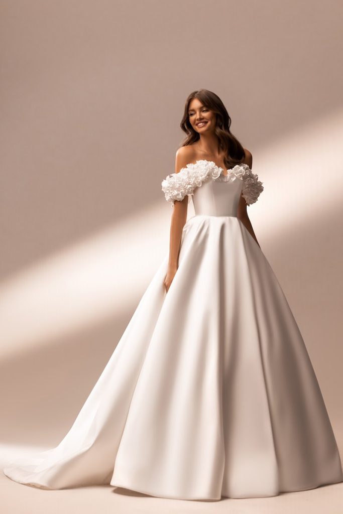 In questa foto un abito da sposa romantico di Katy Corso con gonna ad A, bustino rigido e fiori 3D sullo scollo