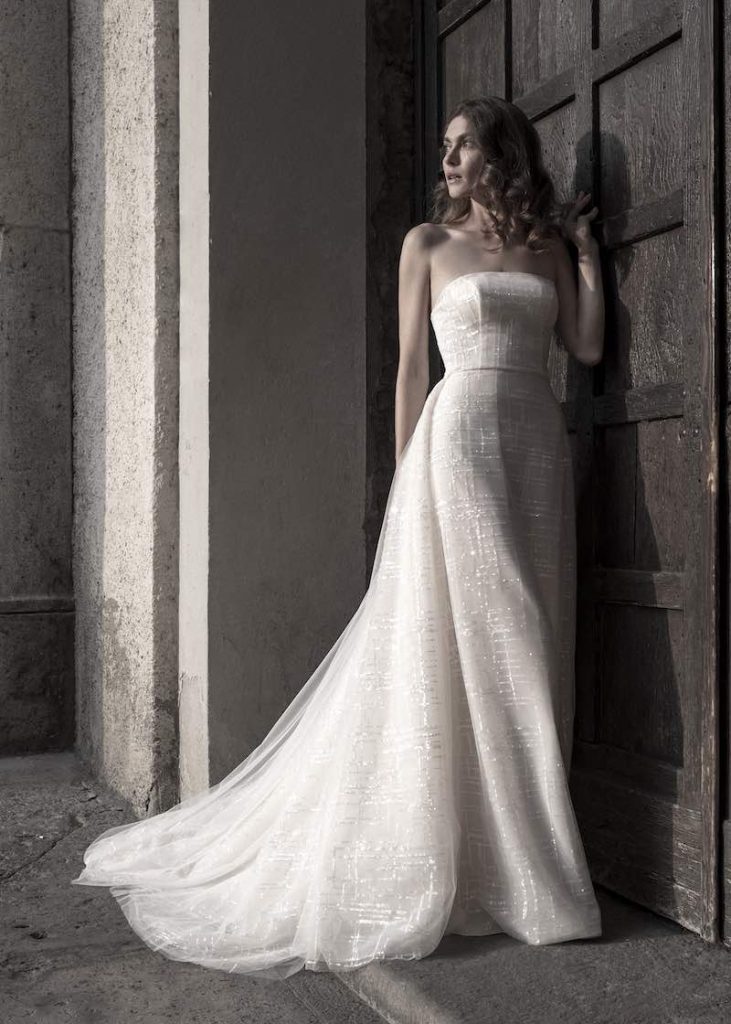 In questa foto una modella posa indossando un abito da sposa a sirena Le Fate Milano in tulle ricamato a righe geometriche con scollatura dritta e sopragonna 