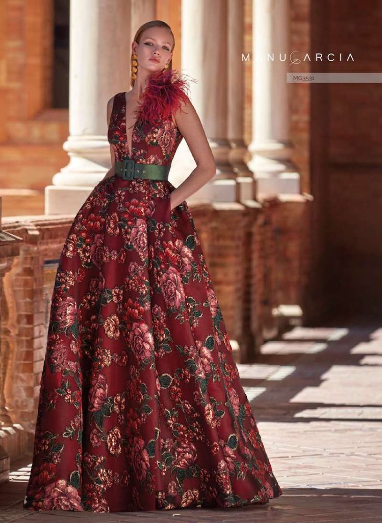 In questa foto una modella indossa un abito da cerimonia lungo e svasato Manu Garçia di colore bordeaux damascata con trama a forma di rose con maxi cintura di colore verde bosco e spilla di piume abbinata in vendita a Palermo presso Magazzini D'Amico