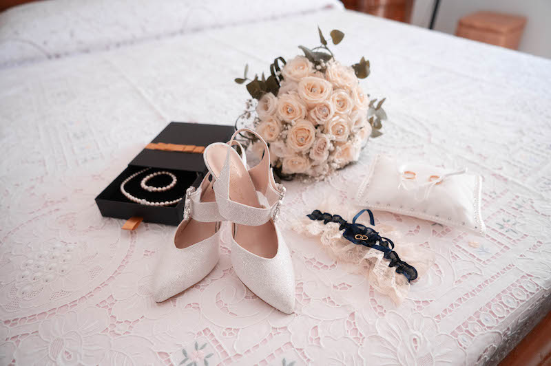 In questa foto di Virgilio Ferrara gli accessori di una sposa (gioielli, scarpe, giarrettiera e il bouquet) e il cuscino con le fedi poggiate su un letto matrimoniale 
