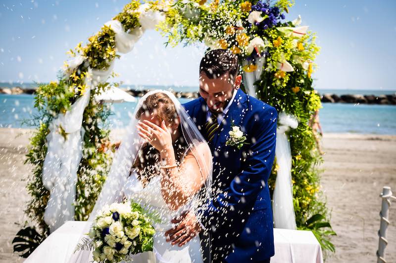 In questa foto due sposi durante il lancio del riso al termine di un matrimonio al Blu Suite Resort sulla spiaggia. Sullo sfondo si intravedono decorazioni floreali di colore giallo e blu