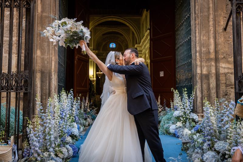 In questa foto due sposi si baciano davanti all'ingresso della chiesa del loro matrimonio allestito con fiori nei toni del blu