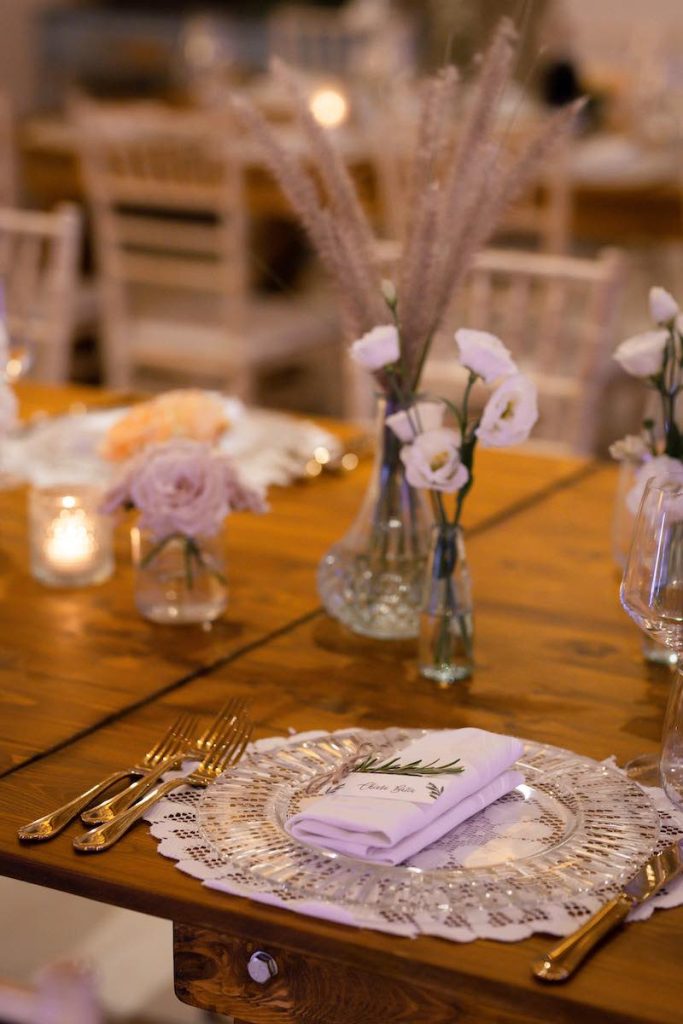 In questa foto il dettaglio di una mise en place boho chic composta da piatto di vetro, un centrino come sottopiatto, posate d'argento e piccoli vasetti di vetro con rose e fiori di colore bianco 