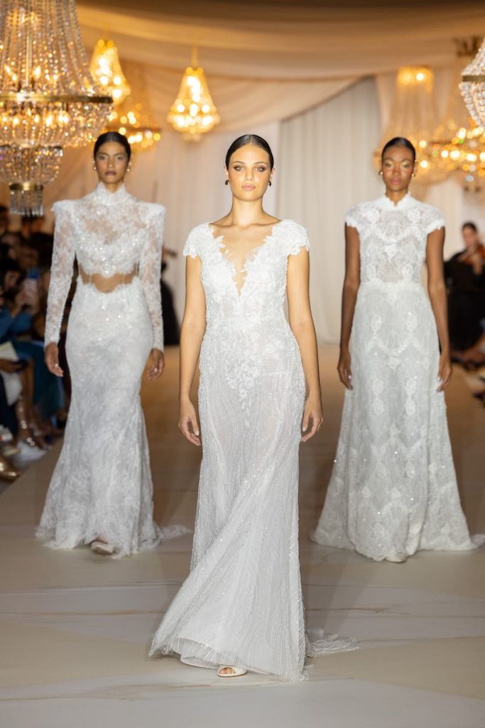 In questa foto tre modelle sfilano in passerella con tre abiti da sposa modello sirena con ricami di pizzo e dettagli gioiello della collezione sposa Atelier Emé 2024