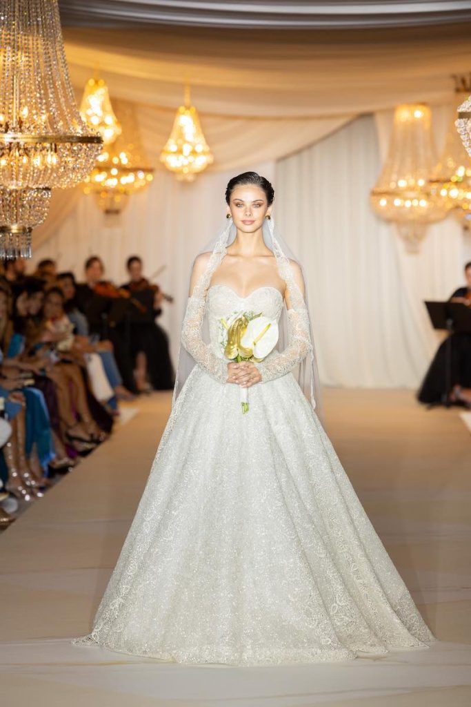 In questa foto una modella durante la sfilata di Atelier Emé 2024 con un abito da sposa ad A, in pizzo con scollo a cuore e velo