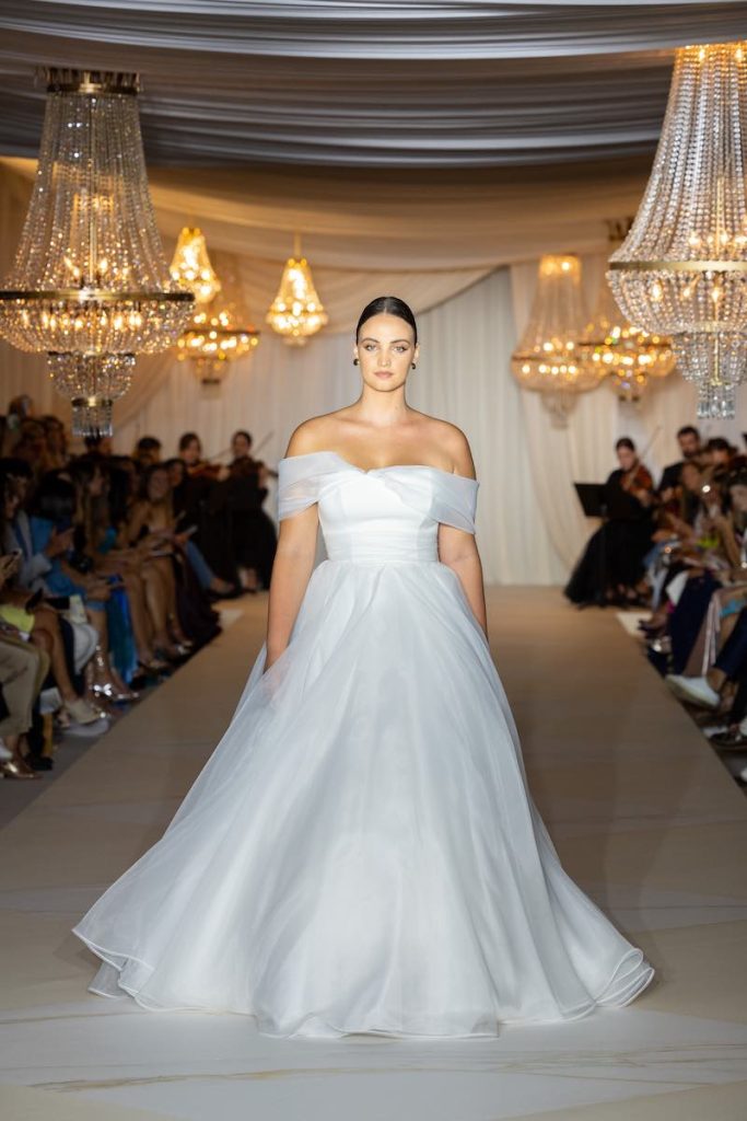 In questa foto una modella sfila in passerella con un abito da sposa modello ad A di organza con scollo off-shoulder della collazione 2024 Atelier Emé