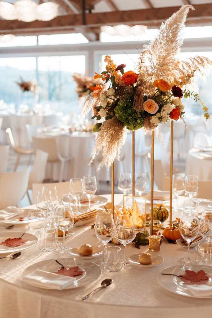In questa foto un centrotavola a tema autunno con pampas, fiori di colore rosa e arancione e zucche