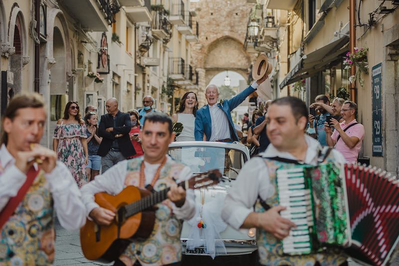 In questa foto di Alessandro Tondo due sposi inglesi a bordo di una Cinquecento di colore bianco festeggiano il loro matrimonio mentre percorrono una via di Taormina. Davanti a loro tre musicisti con una fisarmonica, una chitarra e un flauto