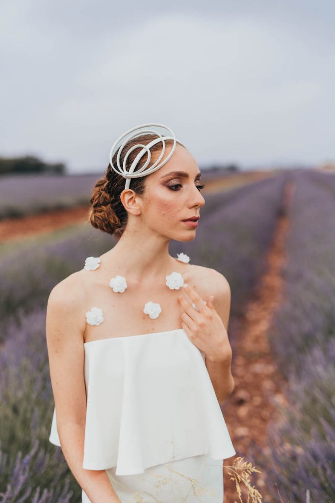 In questa foto una modella il cui look è stato curato dalla Bridal Stylist Giulia Solli posa in un campo di lavanda in Provenza indossando un abito da sposa con scollatura dritta decorata con fiori 3D su tulle illusion e un cerchietto con motivi ellittici