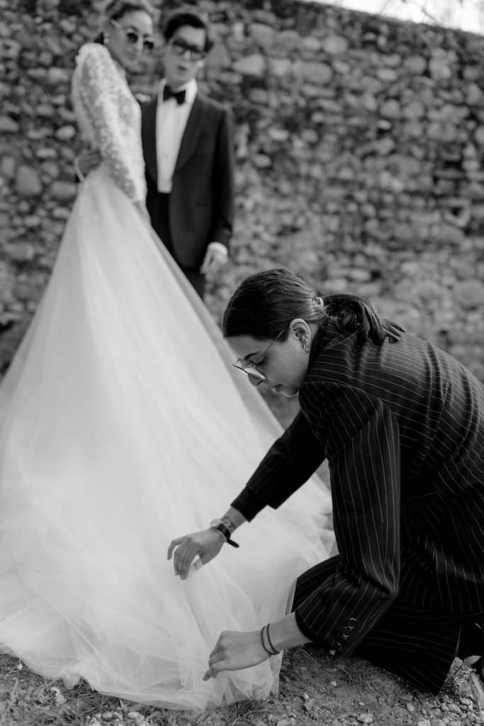 In questa foto in bianco e nero la Bridal Stylist Giulia Solli chinata in basso sistema lo strascico in tulle di un abito su una sposa che abbraccia il suo sposo durante lo shooting di nozze