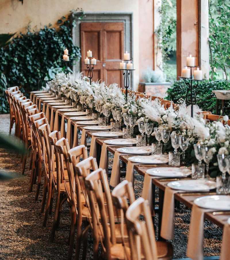 In questa foto un tavolo imperiale inquadrato in obliquo, allestito dalla Wedding Planner Romina Pecorari di RP Wedding & Events con runner di fiori di colore bianco e sedie in legno