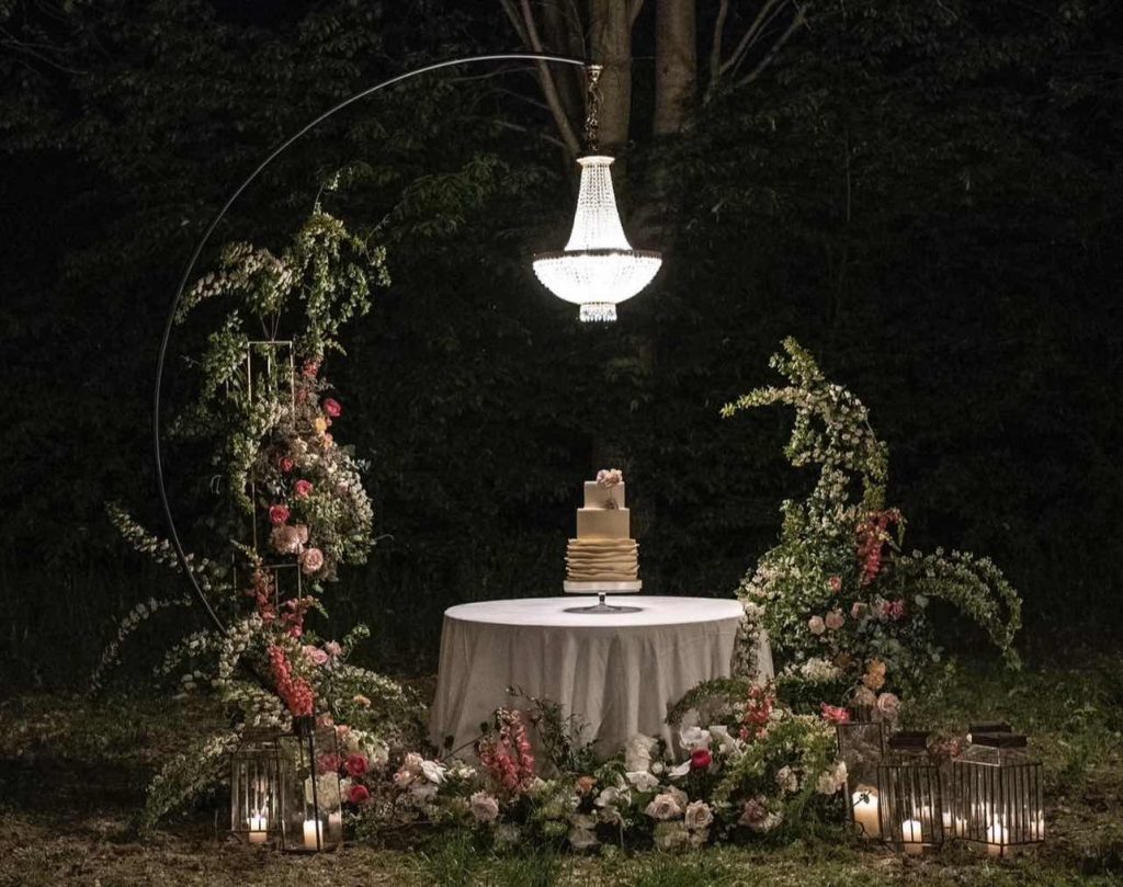 In questa foto il tavolo della torta nuziale allestito dalla Wedding Planner Romina Pecorari di RP Wedding & Events con fiori di colore rosa e bianco su una struttura semicircolare con lampadario Chandelier centrale 
