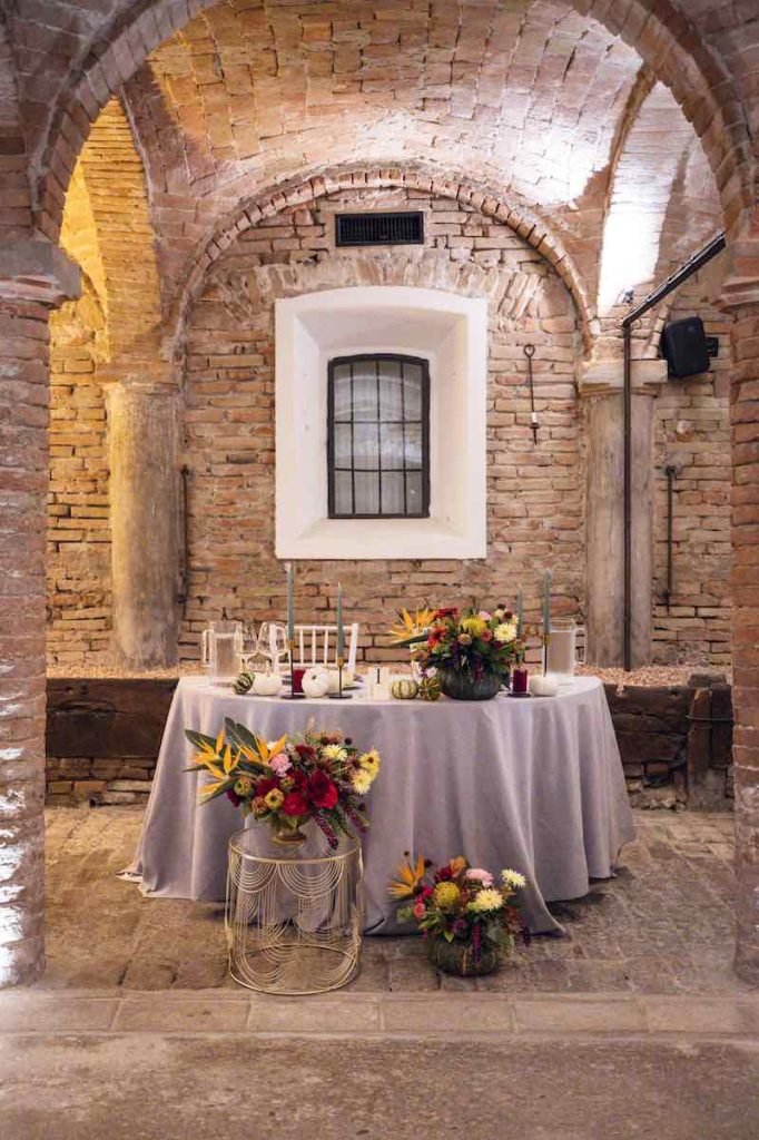 In questa foto il tavolo degli sposi Silvia e Alessandro sotto ad un arco in pietra decorato da OttoQuattro Eventi con tavoli in ferro di colore oro con piano in vetro e composizioni di rose, anthurium, sterlizia, nutan, garofani disposti all'interno di zucche