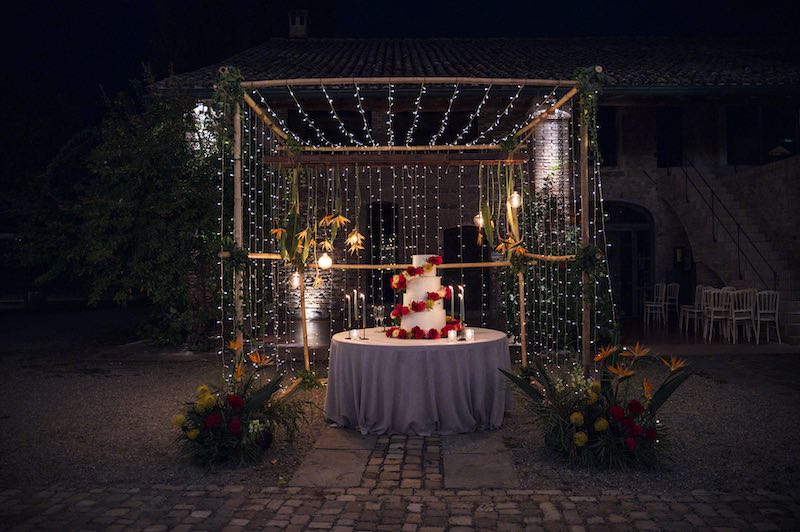 In questa foto lo spazio allestito per il taglio torta del matrimonio di Silvia e Alessandro decorato con un arco di bamboo di piccole luci e composizioni di fiori