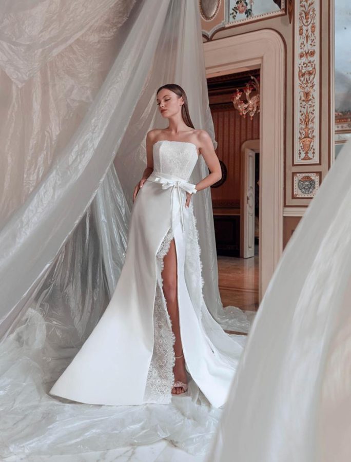 In questa foto una modella posa indossando un abito da sposa a sirena con spacco laterale, inserti in pizzo e fiocco in seta in vita di Rosanna Perrone 