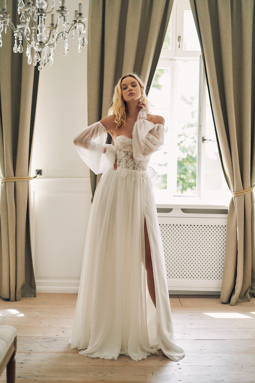 In questa foto una modella in posa con un abito da sposa scivolato 2024 di Annais Bridal: il vestito ha uno spacco sulla gonna in organza e le maniche balloon rimovibili