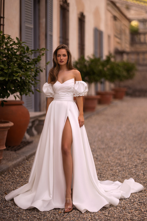 In questa foto una modella indossa un vestito da sposa con maniche a palloncino rimovibili, fascia ton sur ton sotto il seno, scollatura a cuore e profondo spacco sulla coscia. L'abito è di Aria Bride 2024 